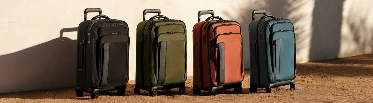 Four Briggs & Riley ZDX suitcases in black, green, orange, and blue in the sun against a white wall