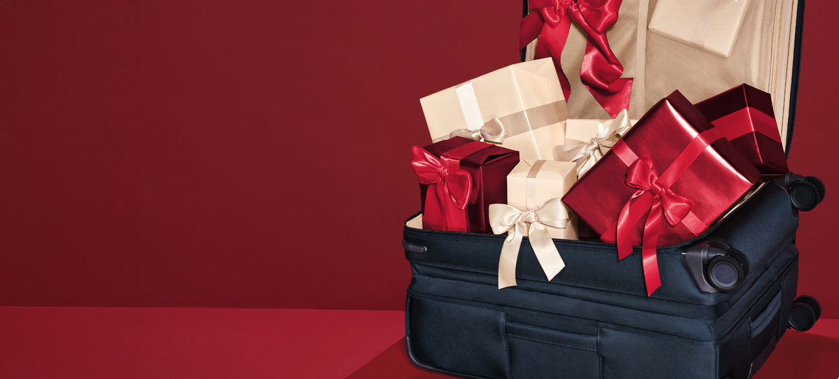 An open navy blue suitcase on a red surface against a dark red background, filled with wrapped gift boxes in red, cream, and brown with satin ribbons.