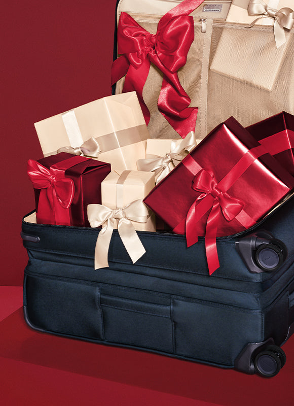 A dark navy blue rolling suitcase filled with wrapped gift boxes in cream and red, tied with large satin bows, set against a deep red background.