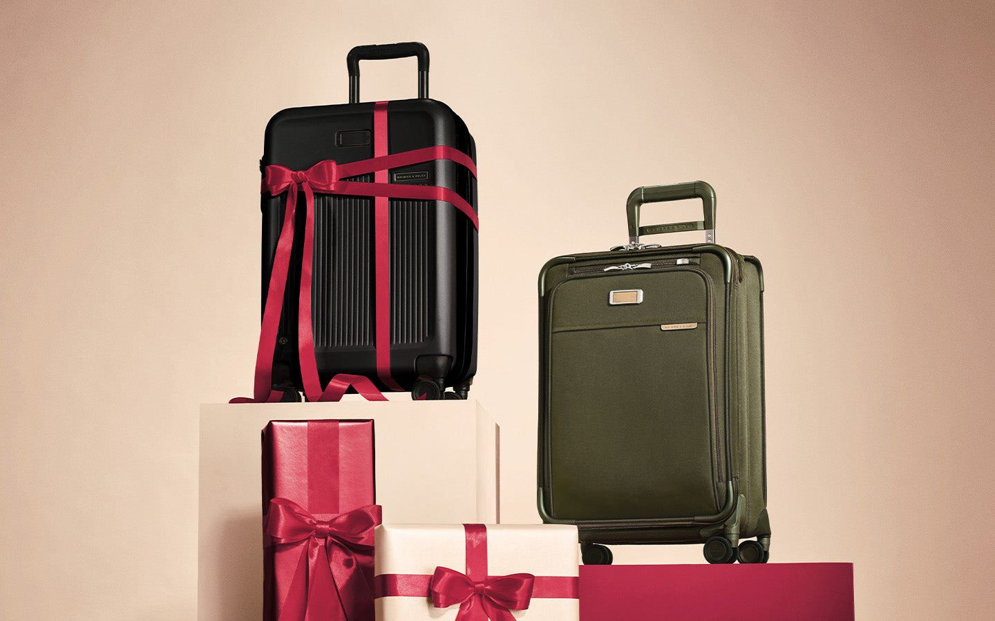 Two hard-shell suitcases displayed on gift boxes against a warm beige background. The left suitcase is black with a dark red ribbon wrapped around it, and the right suitcase is olive green with a small metallic brand plaque.