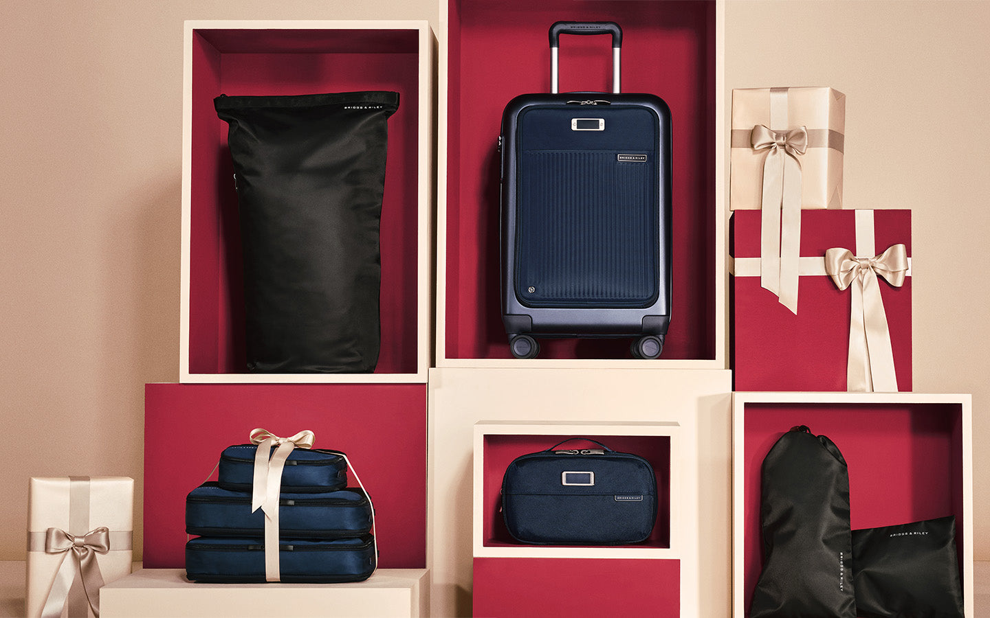 A travel luggage set displayed in a styled studio setup with cream-colored display boxes and red backdrops. The set includes a hard-shell carry-on suitcase, a soft-sided duffel bag, a stack of matching checked luggage pieces, and a small zip pouch, with some pieces wrapped in gift paper and ribbon.