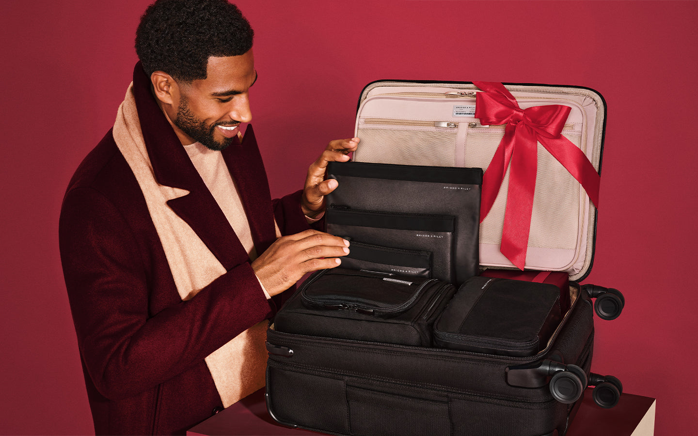 A man in a burgundy blazer and cream scarf opens a black suitcase, revealing organized black travel gear inside, with a large red ribbon tied around the open suitcase against a solid red background.